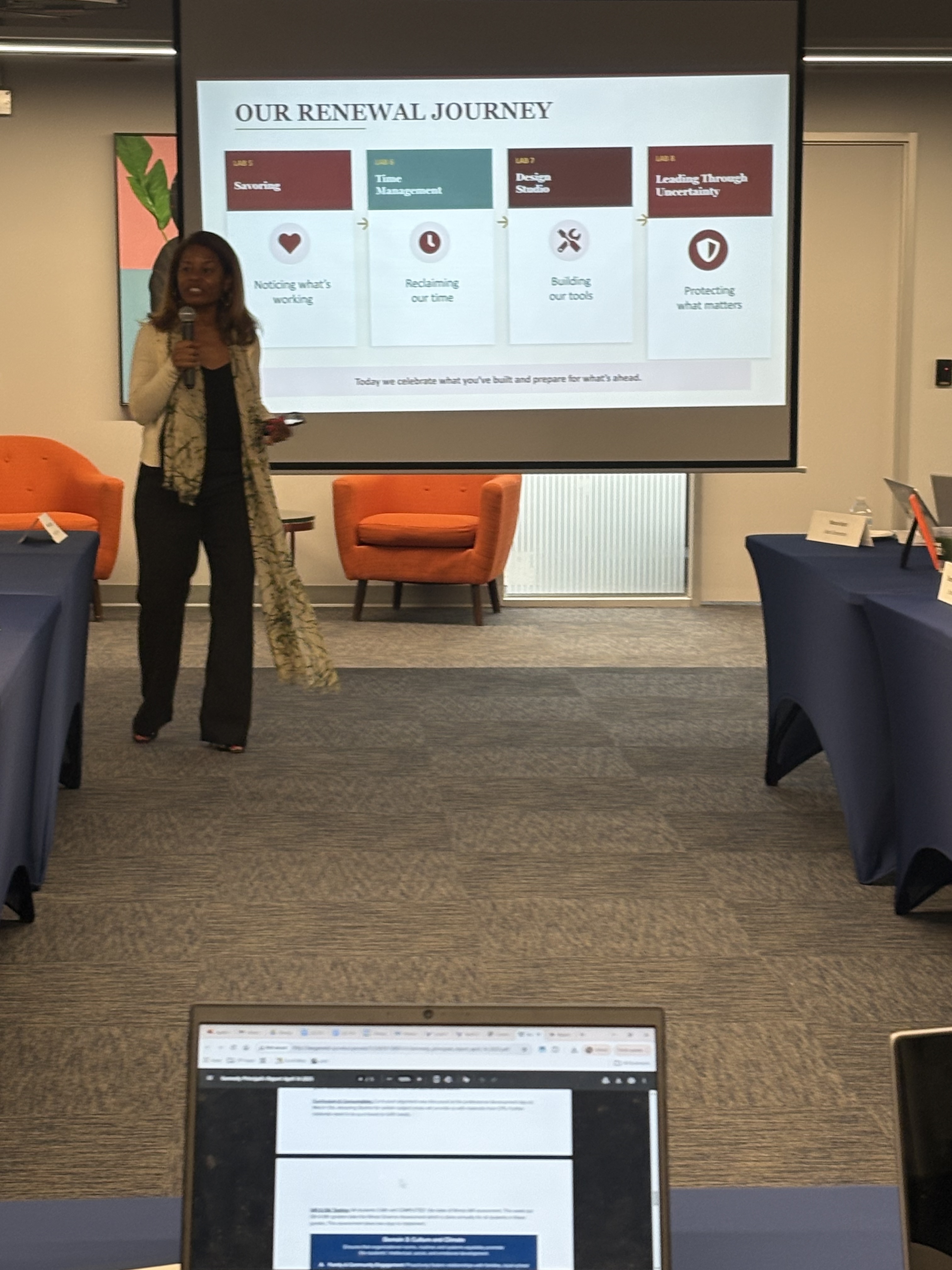 Dr. Peggie Burnett-Wise stands at the front of a learning lab holding a microphone. Behind her is a projected slide titled Our Renewal Journey showing four labs: Savoring, Time Management, Design Studio, and Leading Through Uncertainty.
