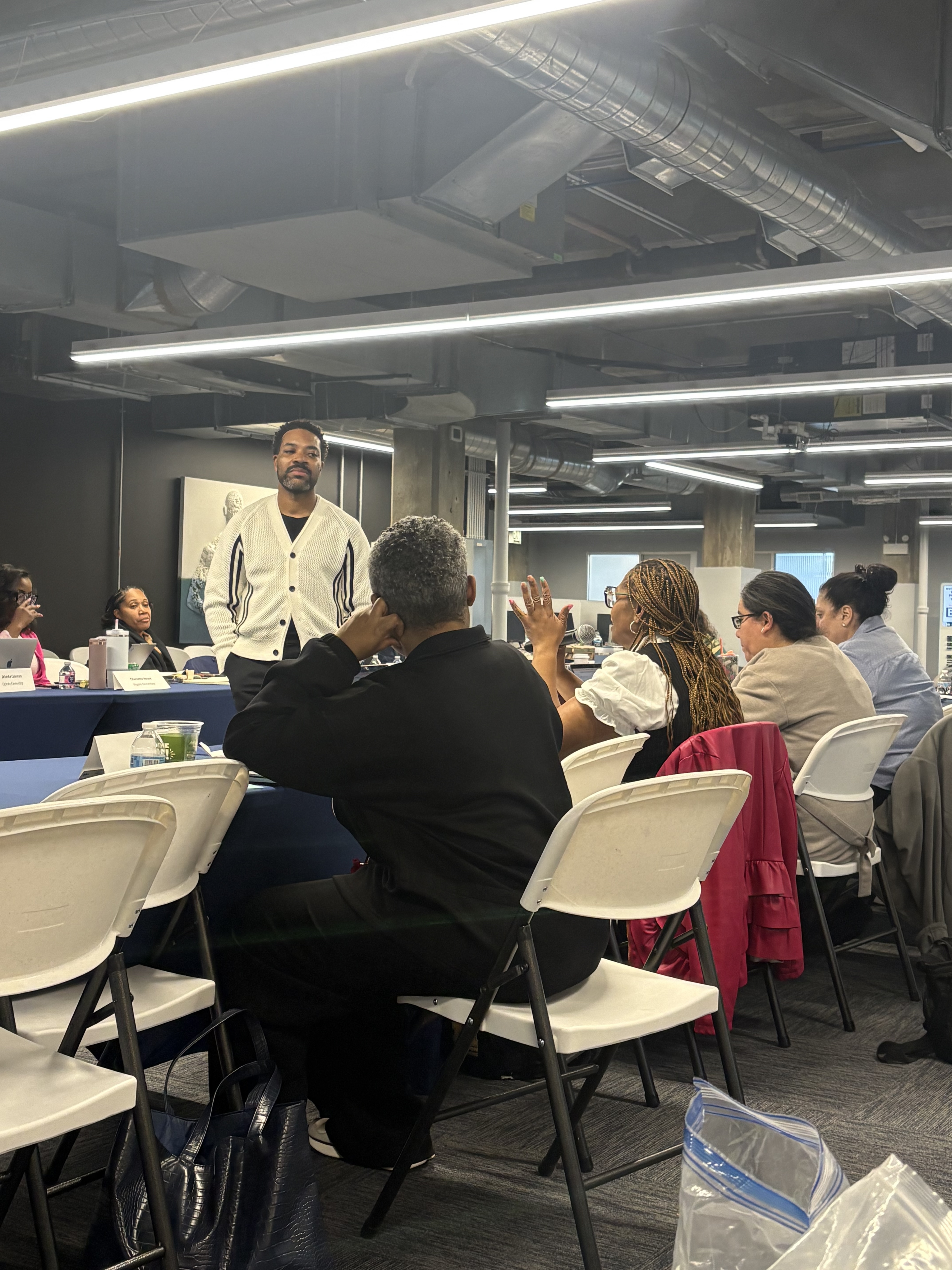 A coach in a white cardigan stands at a table while two principals work through an idea together. One principal is gesturing with both hands; the conversation is clearly active.