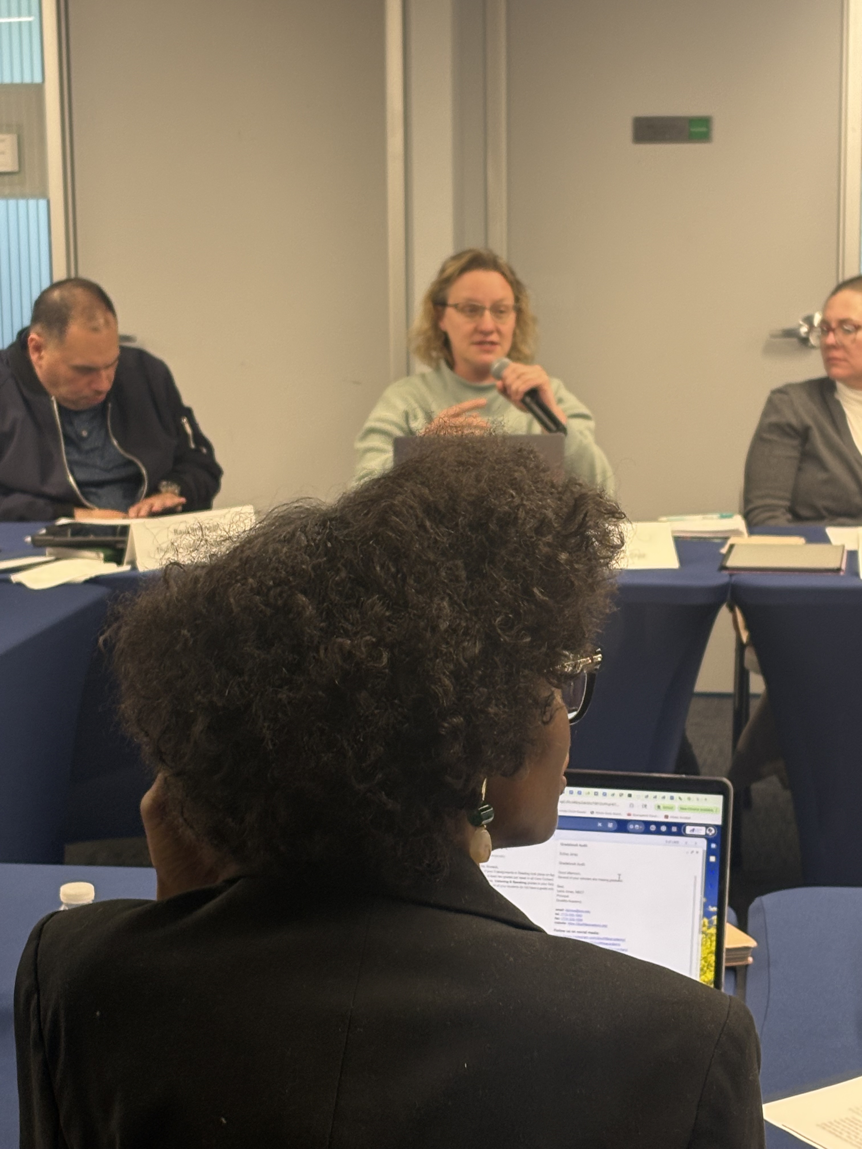 A principal speaks into a handheld microphone, gesturing as she addresses her colleagues. Two other principals are listening in the foreground.